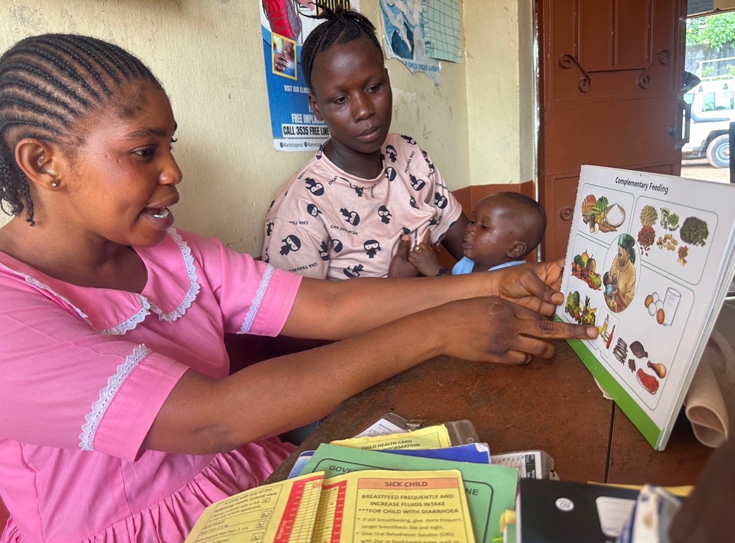 A Sierra Leonean nurse sits next to a mother holding a baby. The nurse points to a chart about complementary feeding.
