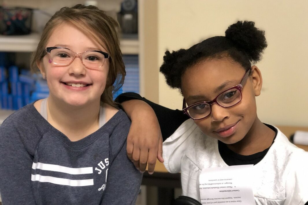 Two girls sitting closely, smiling at the camera, both wearing glasses.