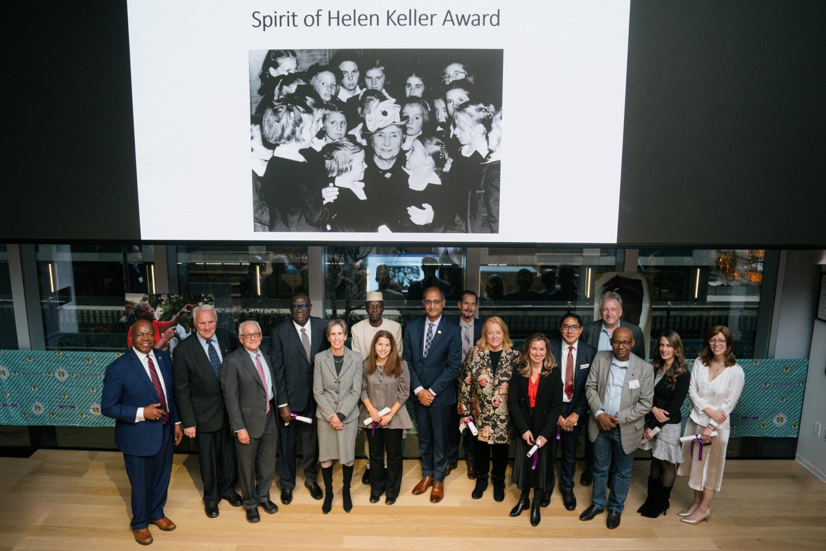 A large group of people stand under a projector screen. On the screen is a black and white photo of Helen Keller with a group of girls. Text at the top of the screen reads "Spirit of Helen Keller Award."
