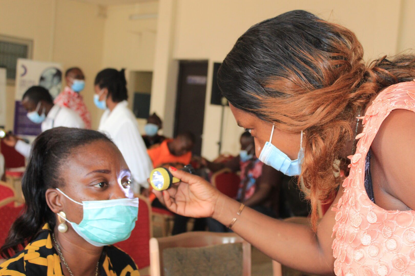 A female community health worker in Cameroon shines a light in a woman's eye as part of an eye exam. 