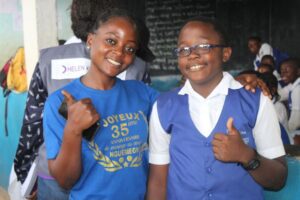 A young woman and an adolescent boy stand next to each other in a classroom in Cameroon. They are smiling at the camera and each giving a thumbs up.