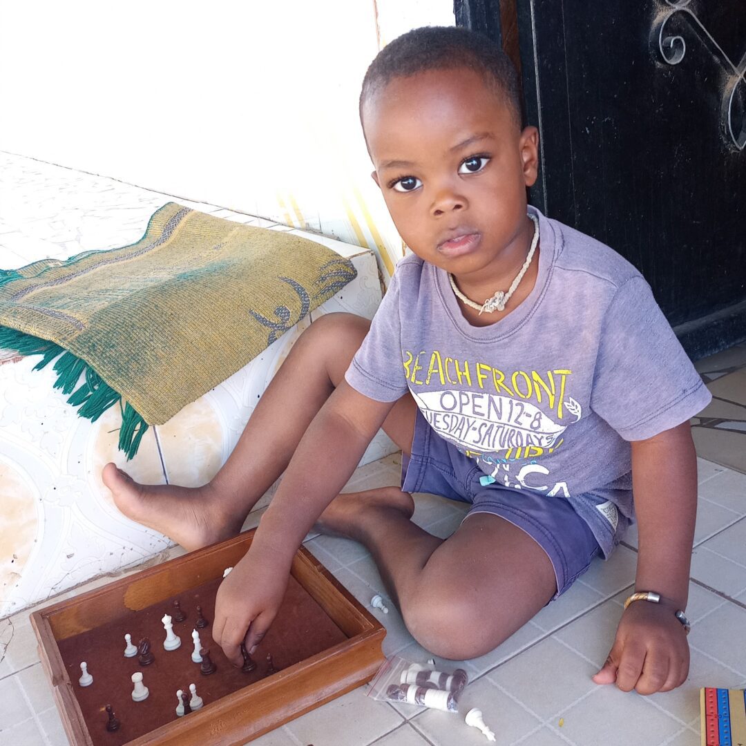 A young Senegalese boy plays with a chess set while sitting on a tile floor.