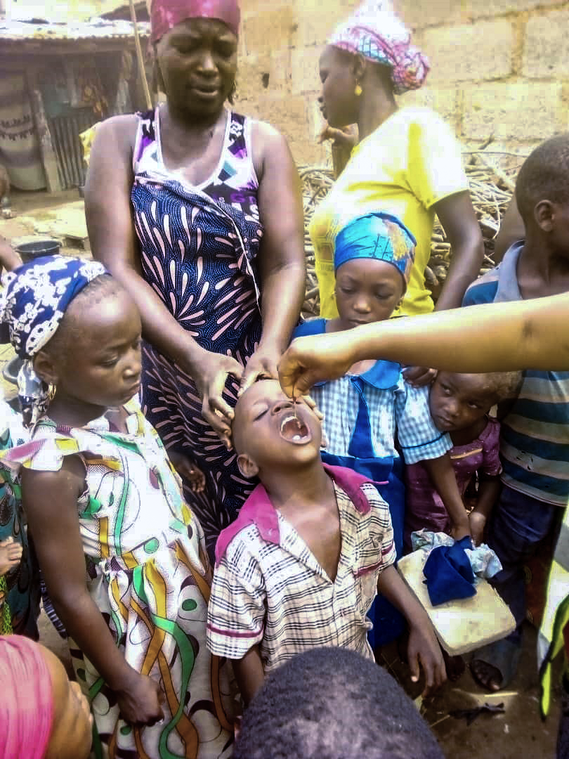 A young Nigerian boy receives a dose of vitamin A with his mother standing behind him. His siblings stand around him.