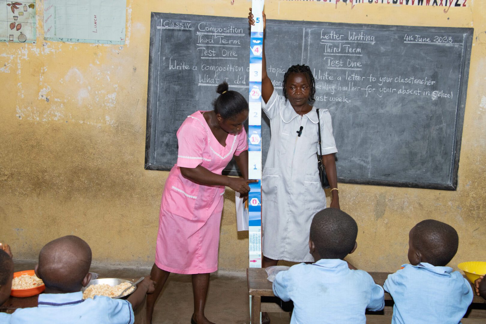 A female healthcare worker and another woman hold up a dose pole, a large measuring stick divided into sections, in front of a classroom of students in Sierra Leone. The healthcare worker points out one of the sections of the dose pole.