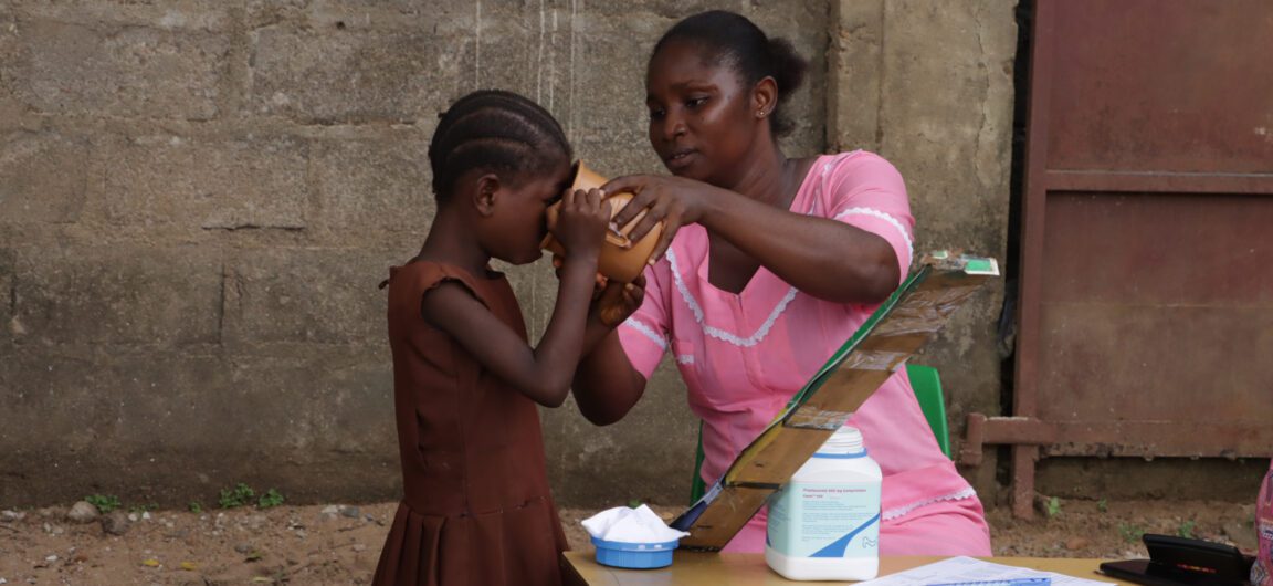 A female healthcare worker in Sierra Leone helps a child take a drink of water. There is a large bottle of medication on the table in front of her.
