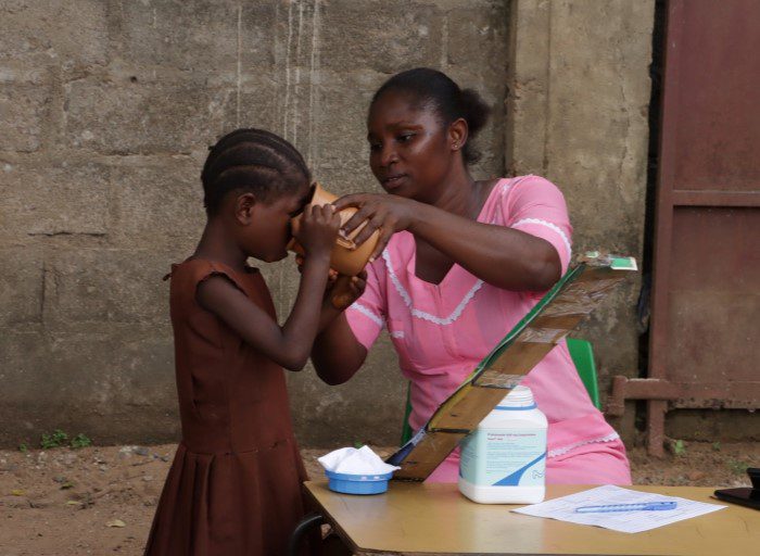 A female healthcare worker in Sierra Leone helps a child take a drink of water. There is a large bottle of medication on the table in front of her.