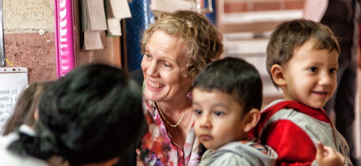 A woman with short, blond hair smiles as she bends down to speak with a group of young children.