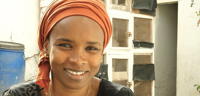 Portrait of a cheerful woman in an orange headscarf smiling outdoors by a white wall with small windows.