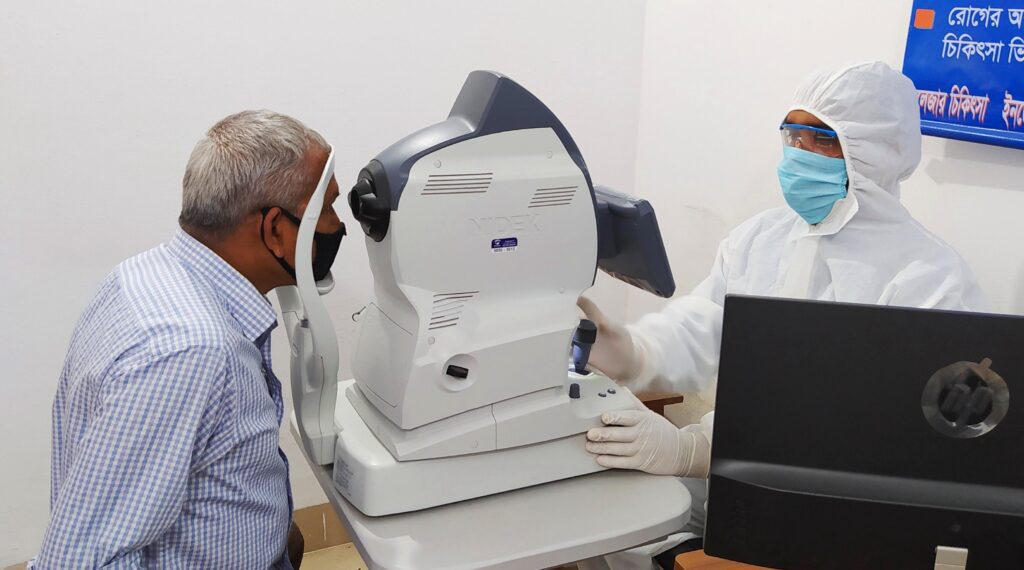 Senior man receiving eye test using advanced ophthalmic equipment while healthcare professional in protective gear operates the machine.