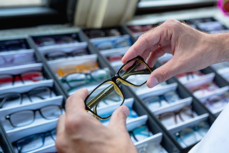 Hands holding and examining a pair of eyeglasses frames over a tray with multiple glasses.