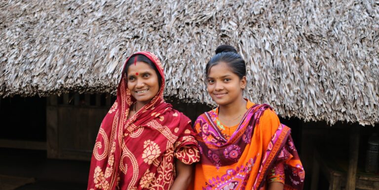 Woman and young girl wearing colorful sarees stand smiling in front of a rural thatched roof home.