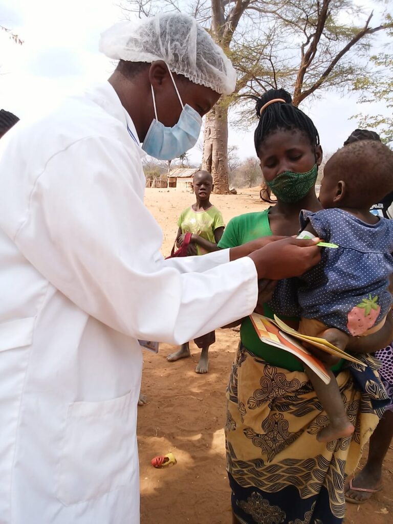 Healthcare professional in protective gear uses MUAC tape to screen young child for malnutrition in rural setting.