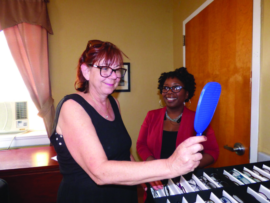 Woman wearing glasses looks at herself in mirror standing next to another woman wearing glasses.