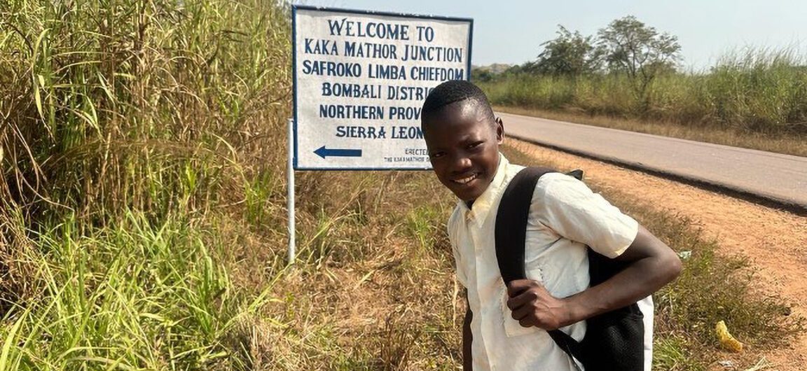A Sierra Leonean boy wearing a backpack stands in front of a road sign.
