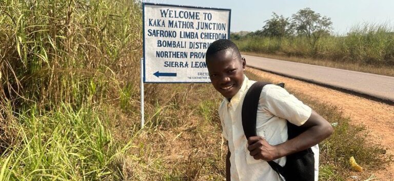 Boy with backpack smiling beside a road sign in a grassy rural area of Sierra Leone.
