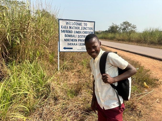 A Sierra Leonean boy wearing a backpack stands in front of a road sign.