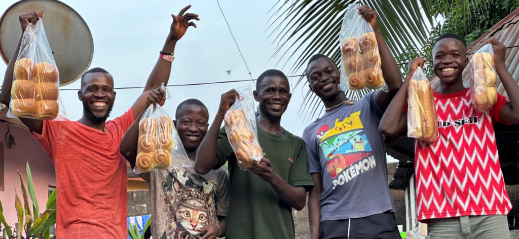 Five young black men hold up bags of bread and rolls. they are standing outside with some buildings and trees in the background.