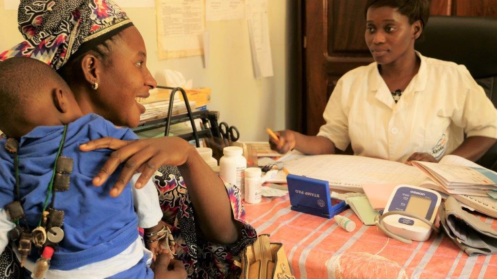 A woman holds her baby and smiles at a nurse in rural clinic setting.
