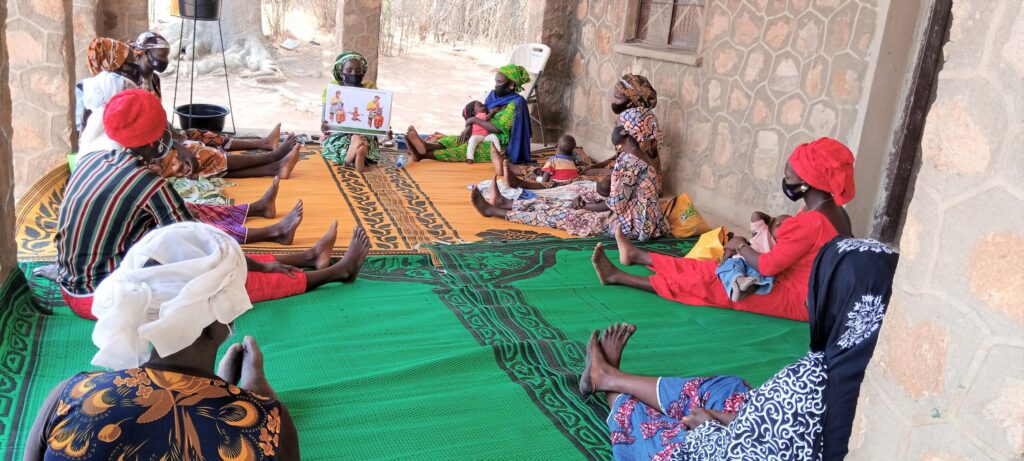 Several women wearing colorful traditional attire sit on green mats inside a rustic building, participating in a health discussion.