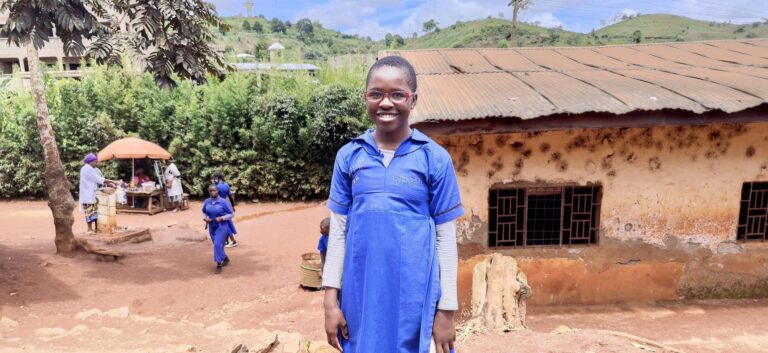 Schoolgirl wearing eyeglasses in a blue uniform stands outside a rustic school building in a rural area