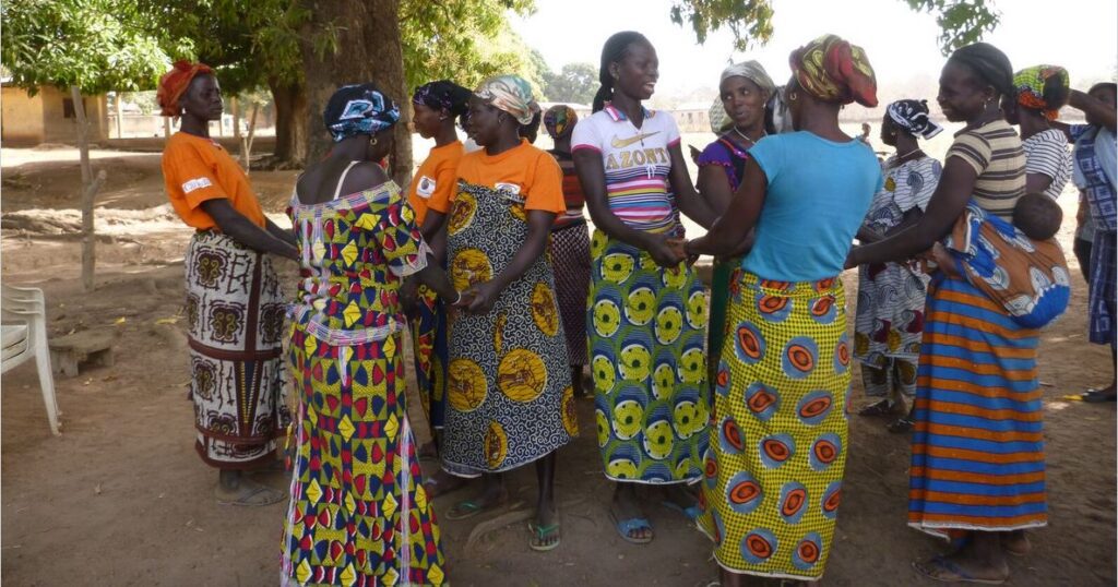 African women wearing vibrant traditional clothing gather outdoors in a circle, engaging in a communal activity in a rural setting.