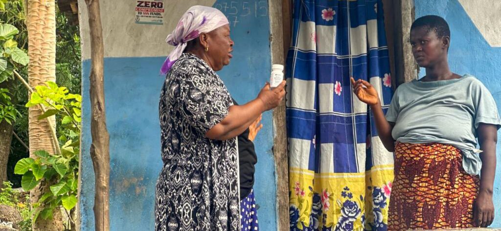 Two Sierra Leonean women stand outside of a house. One is holding a bottle of prenatal vitamins.