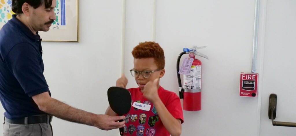 A young American boy gives a double thumbs up as a man holds up a mirror for the child while he is trying on glasses as part of vision care.