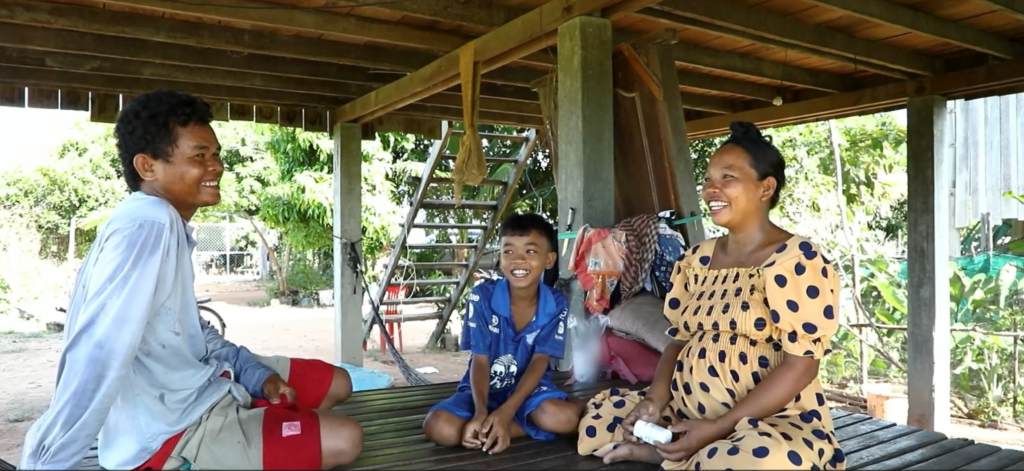 A father, son, and mother sit under their raised house in Cambodia.