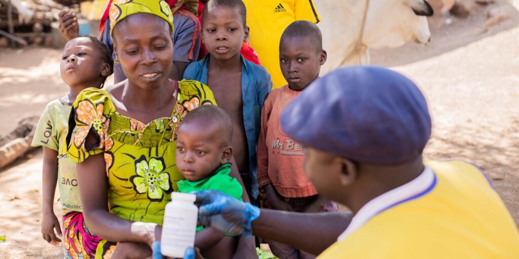 A Cameroonian community health worker shows a bottle if vitamin A to a mother and her children.