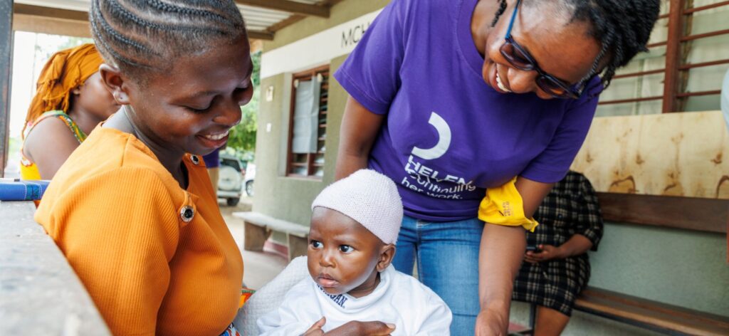 A Kenyan woman holds a small baby on her lap as another woman looks on.