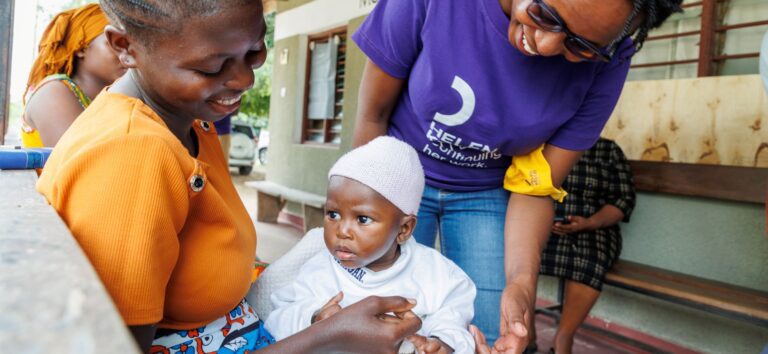 A Kenyan woman holds a small baby on her lap as another woman looks on.