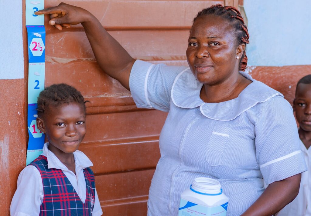 A healthcare worker measures a girl's height. The healthcare worker holds a large bottle of medication.