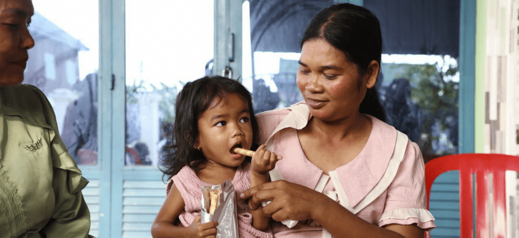 A Cambodian girl eats a nutrient wafer while sitting on her mother's lap.