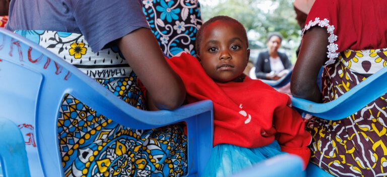 A child in Kenya dressed in a red shirt looks neutrally at the camera while sitting on the lap of their mother.