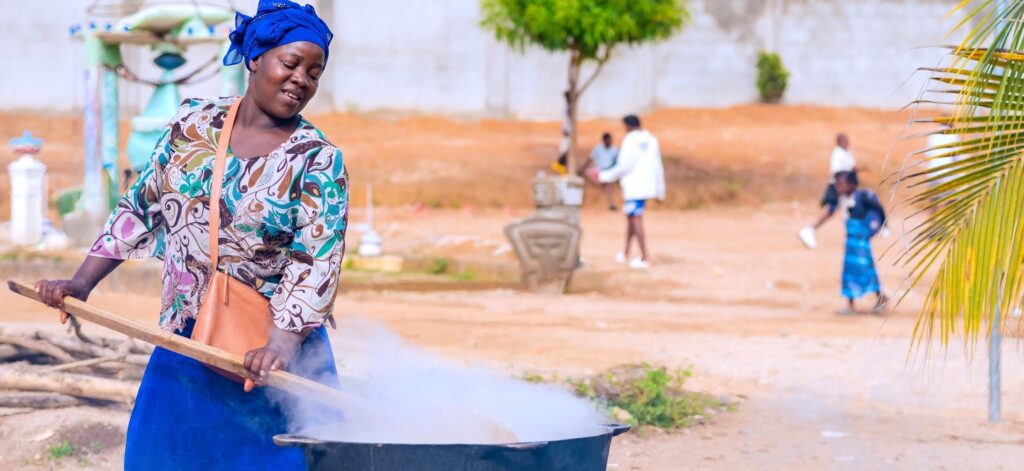 A woman in Sierra Leone stirs a very large pot outside.