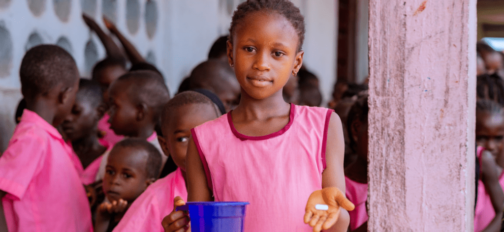 A young girl in Sierra Leone holds out a pill in one hand and a plastic pitcher of water in the other.