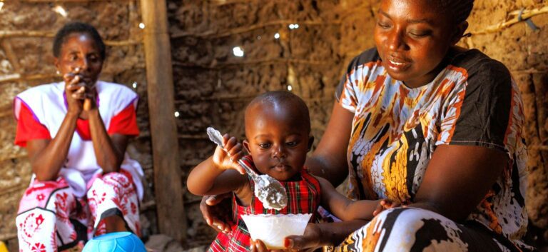 A mother in Kenya holds a small bowl in front of her young child as the child raises as spoon to the bowl. A community health worker looks on in the background.