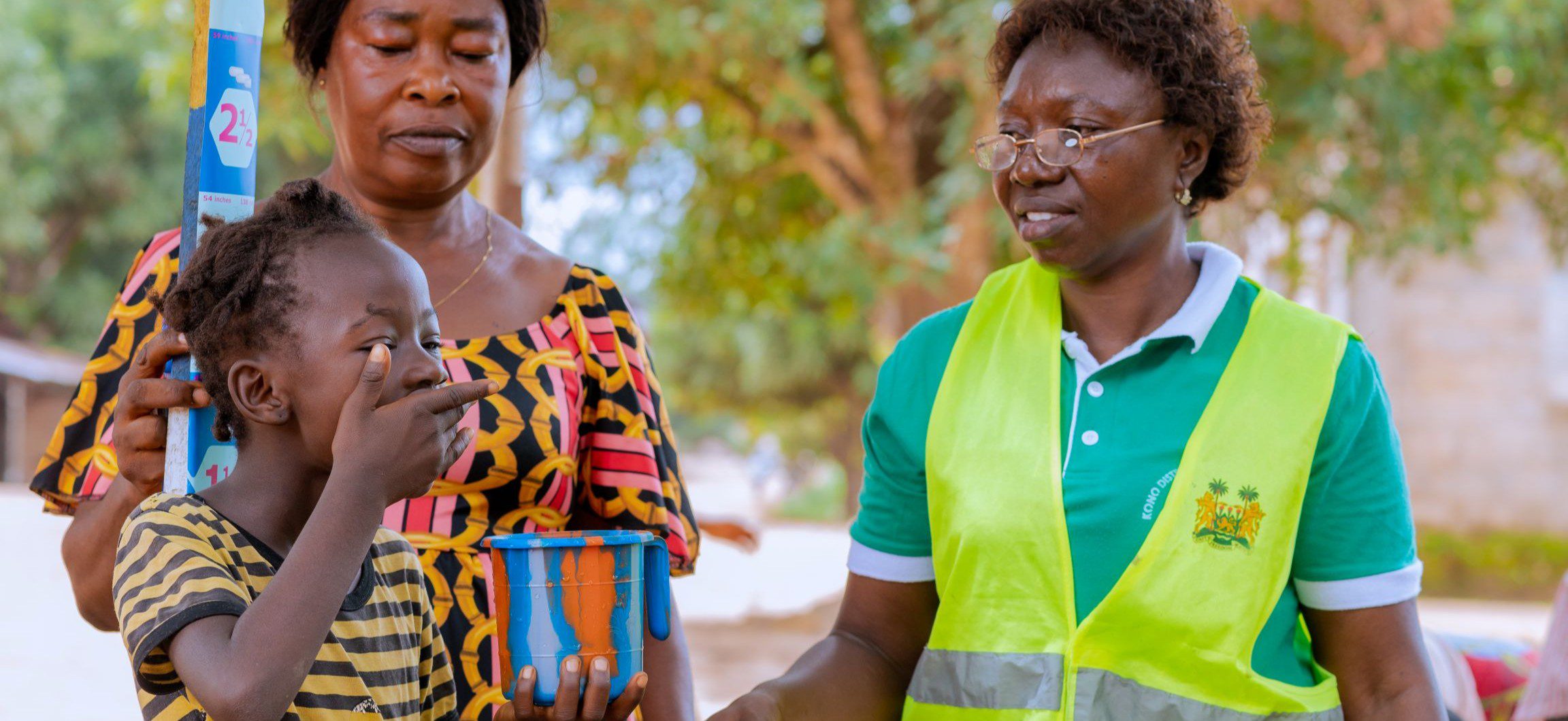 A child in Sierra Leone takes a preventive medication to protect against neglected tropical diseases as two women look on.