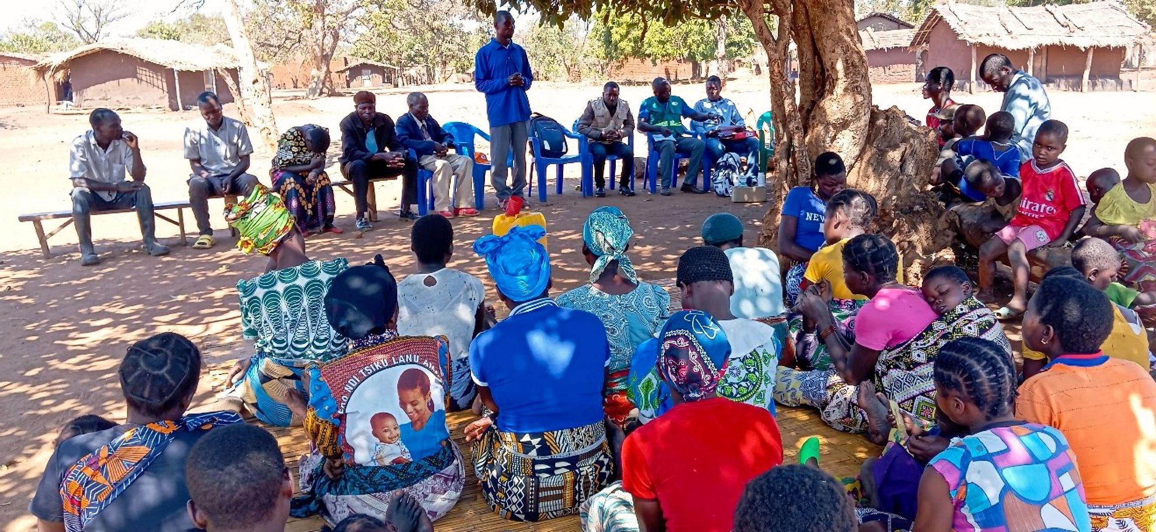 A large group of people sit outside in a community in Mozambique listening to a man speak.