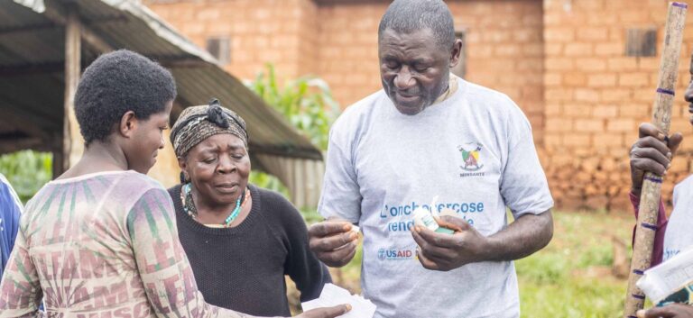 A man in Cameroon hands a pill to a woman as another woman looks on.
