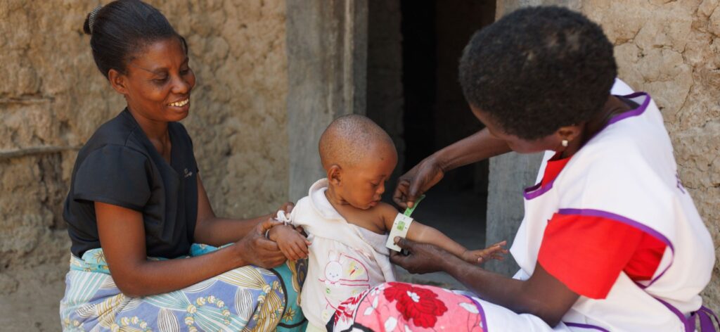 A community health worker in Kenya measures a child's arm circumference to screen for malnutrition as the child's mother looks on.
