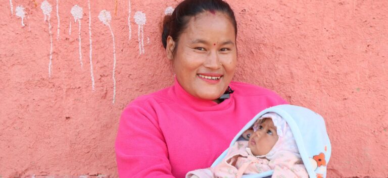 A Nepali woman in pink sweater holding baby wrapped in thick blankets.