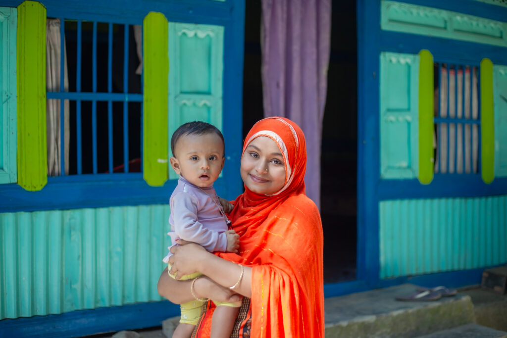 Woman in orange hijab holding baby outside house