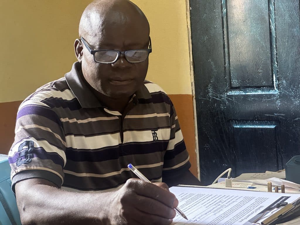 Dominique Veellaah, Eye Health Lead at Moyamba Government Hospital, sitting at a desk indoors with glasses on, holding a pen above a printed document on his desk.