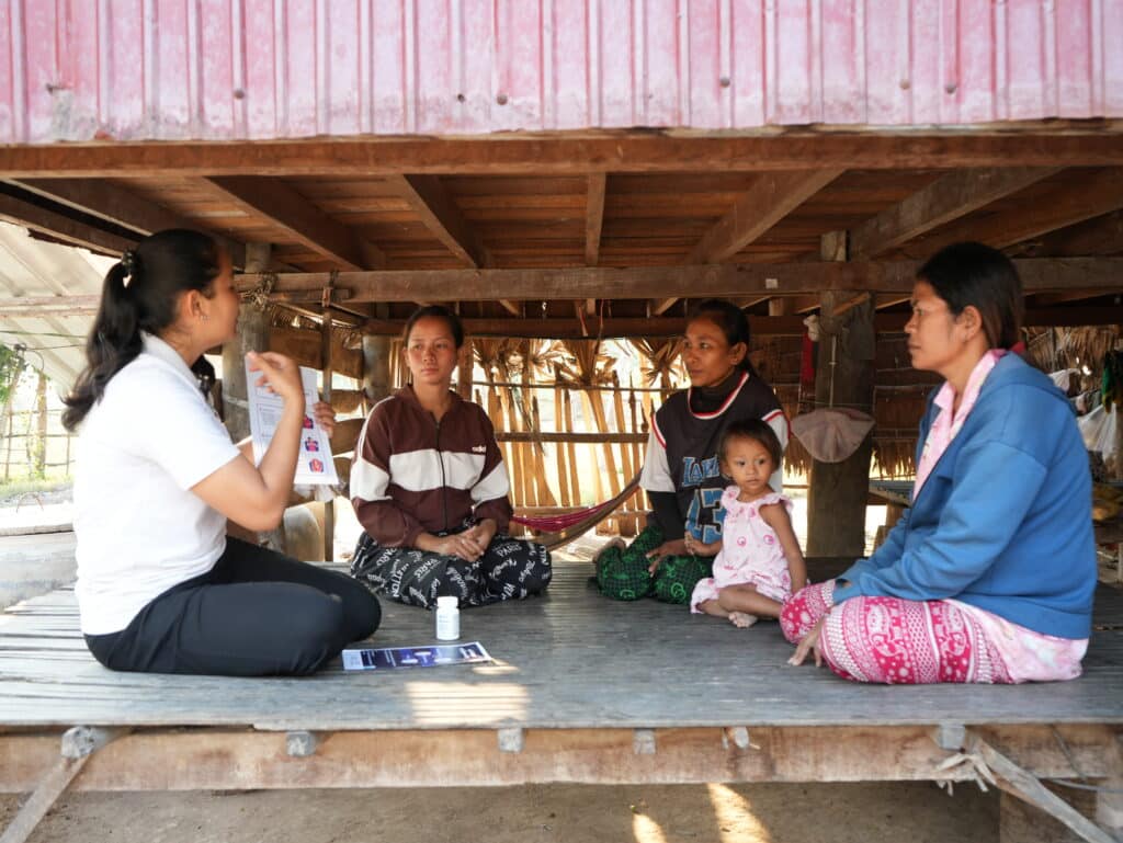 A health worker sitting with a group of women and a young child under a wooden house in Cambodia, holding educational materials about prenatal vitamins.