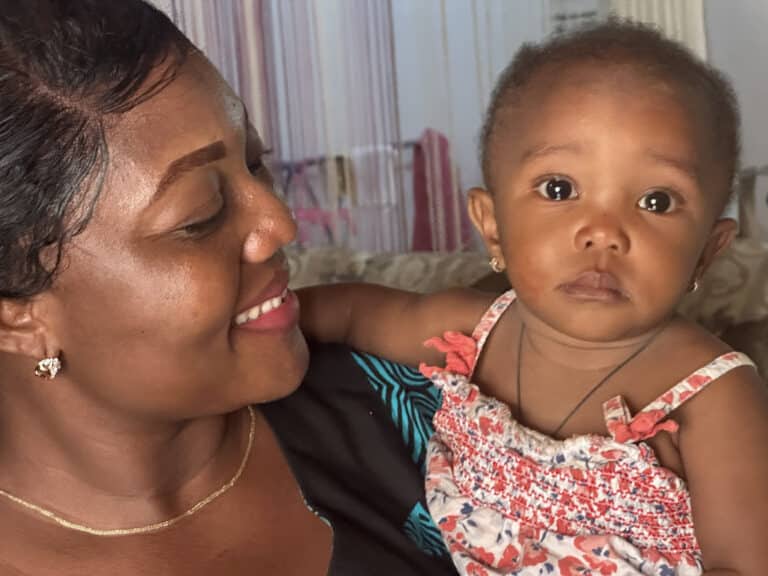 A smiling woman, Koinama, looks lovingly at her 10-month-old daughter, who wears a patterned floral top and looks toward the camera.