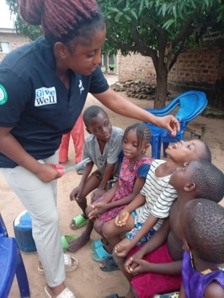 Community health worker Josephine holding a capsule and administering Vitamin A drops to seated children during a rural outreach session under a shade tree.