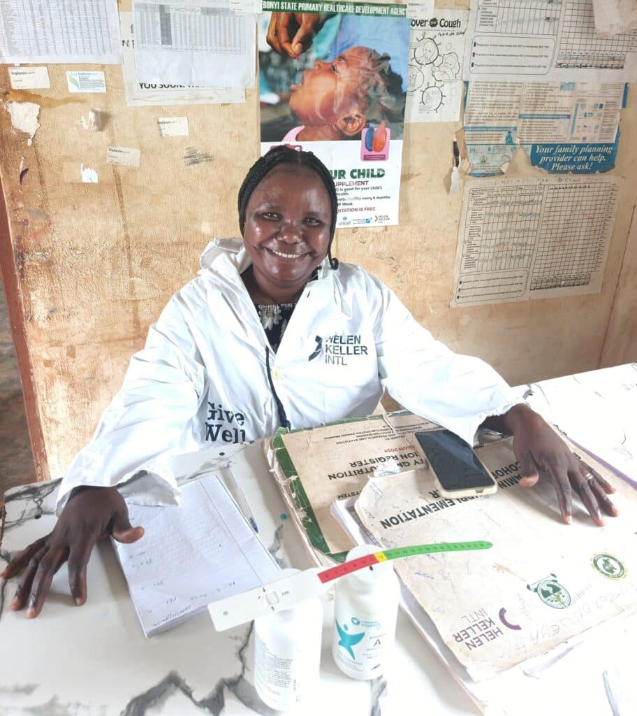 Health worker Nwofe Theresa smiling seated at a desk inside Oguzoronweya Health Centre, wearing a white lab coat with health education posters on the wall behind her.