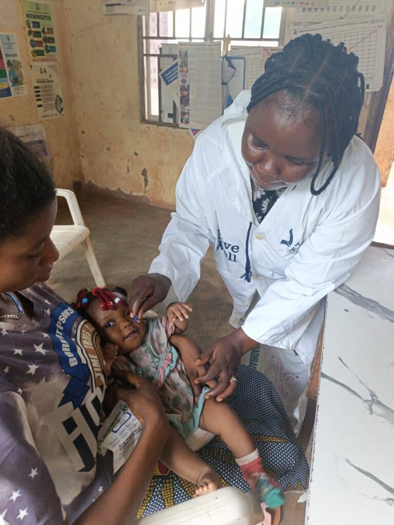 Health worker Theresa leaning over to administer Vitamin A oral drops to a young child seated on a caregiver's lap inside a clinic.