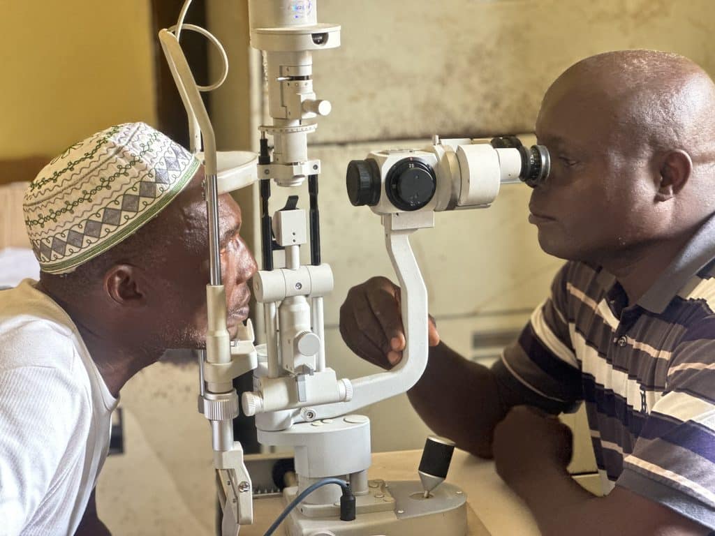 A healthcare provider uses a slit-lamp microscope to examine the eye of a patient wearing an embroidered cap.
